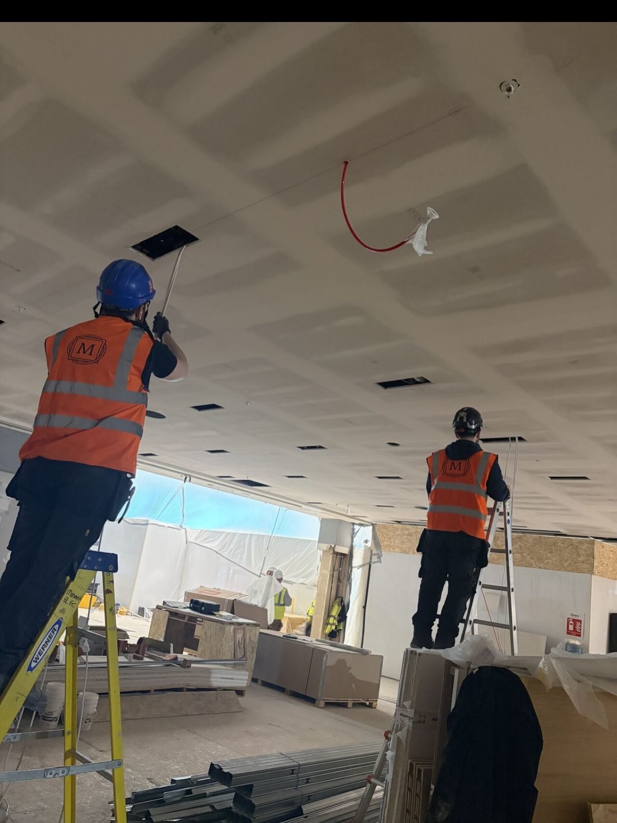 Two electricians in hi-vis on ladders at a ceiling during a fit-out