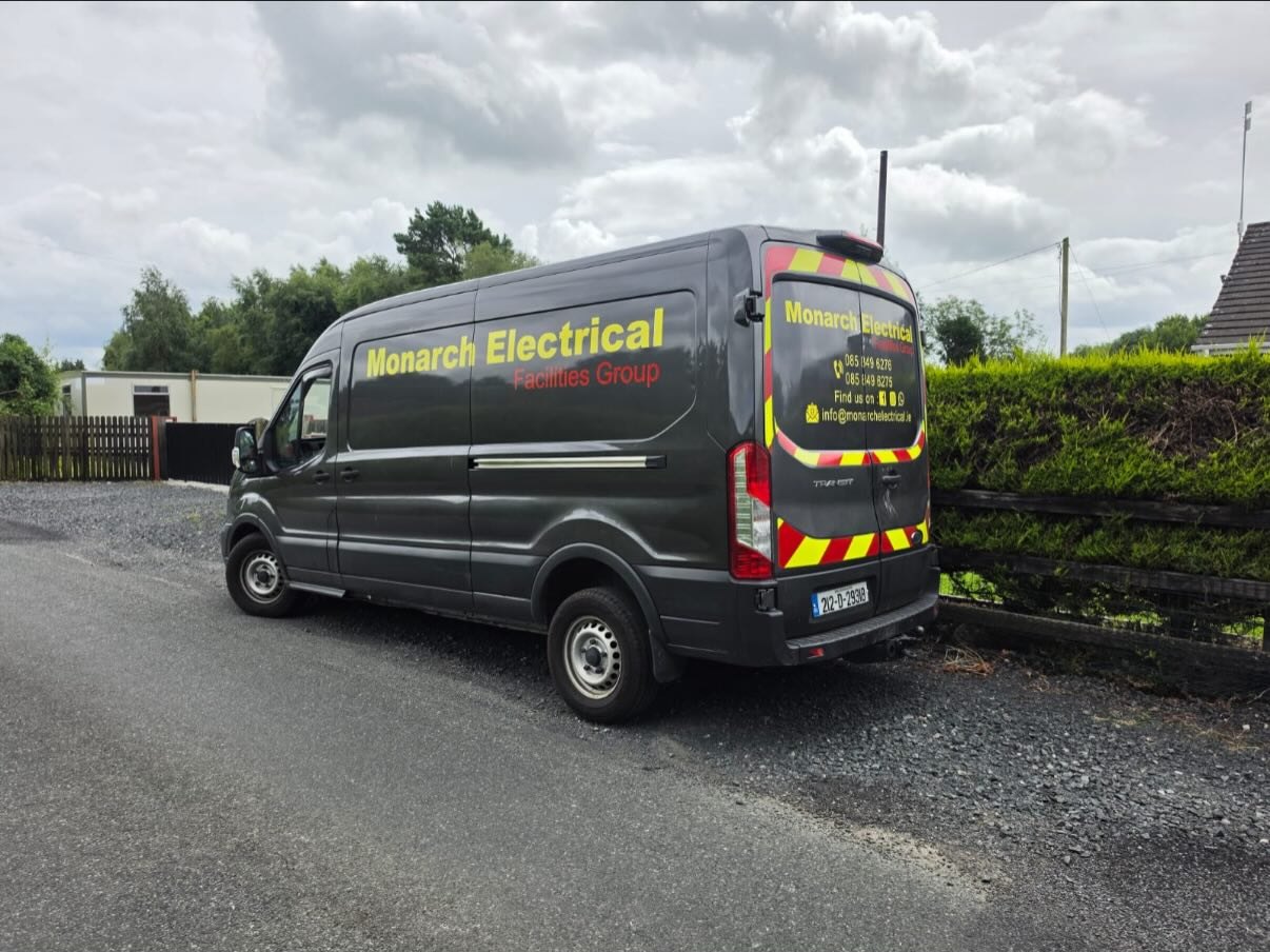 A dark van lettered Monarch Electrical, parked by a hedge